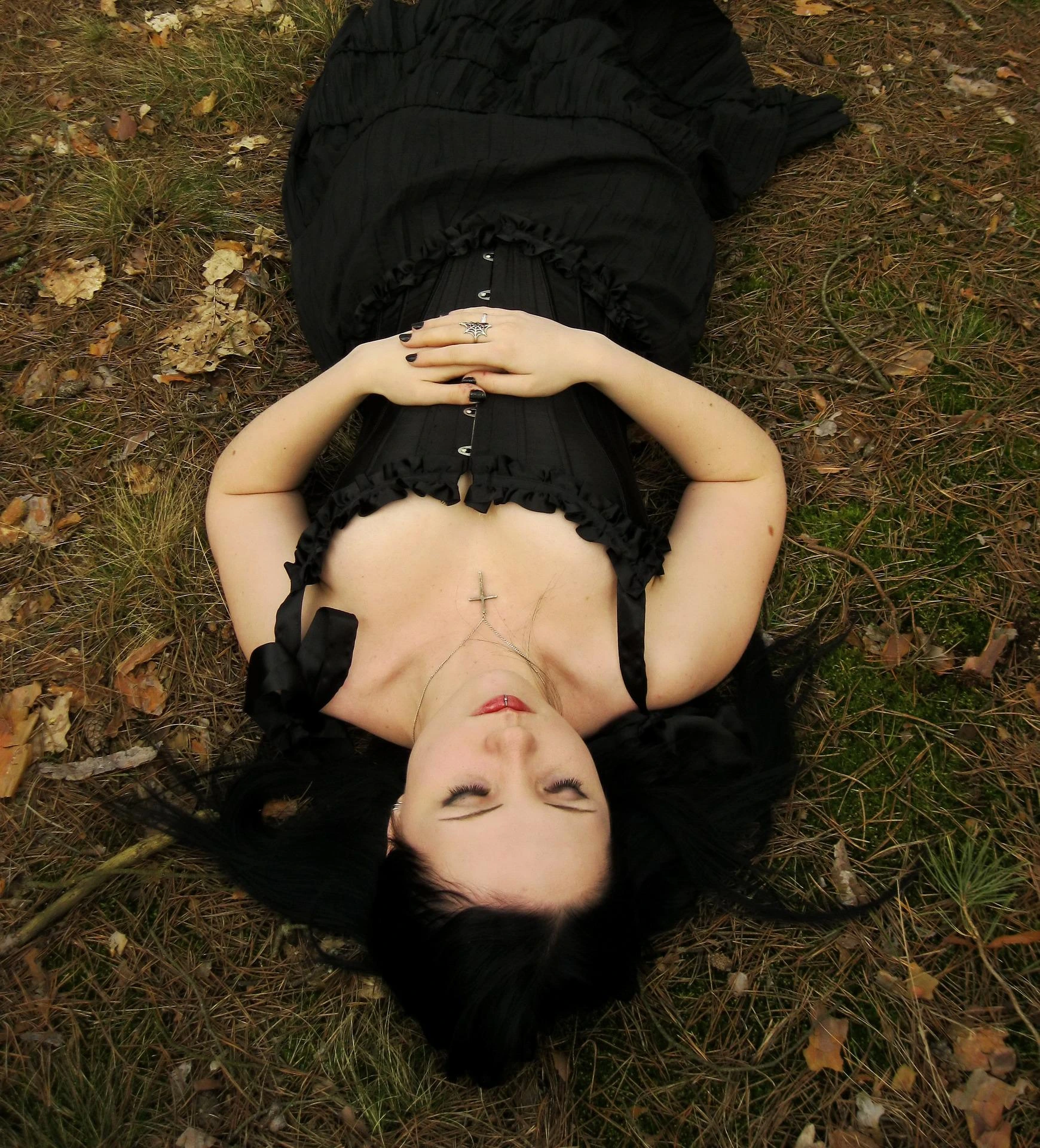 Woman in black dress lying on forest floor with hands folded, eyes closed, surrounded by pine needles.