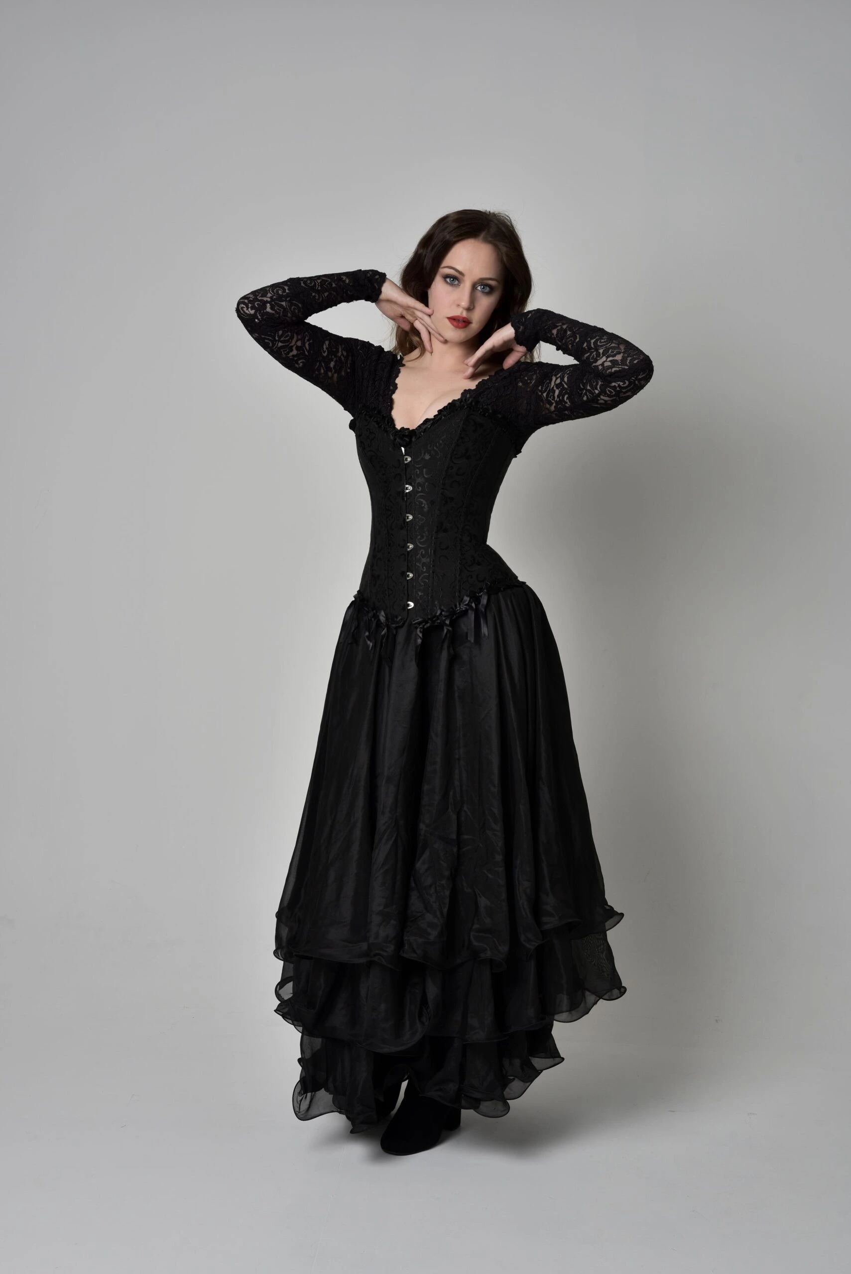 Woman in long black gothic dress and lace sleeves posing against white studio background.
