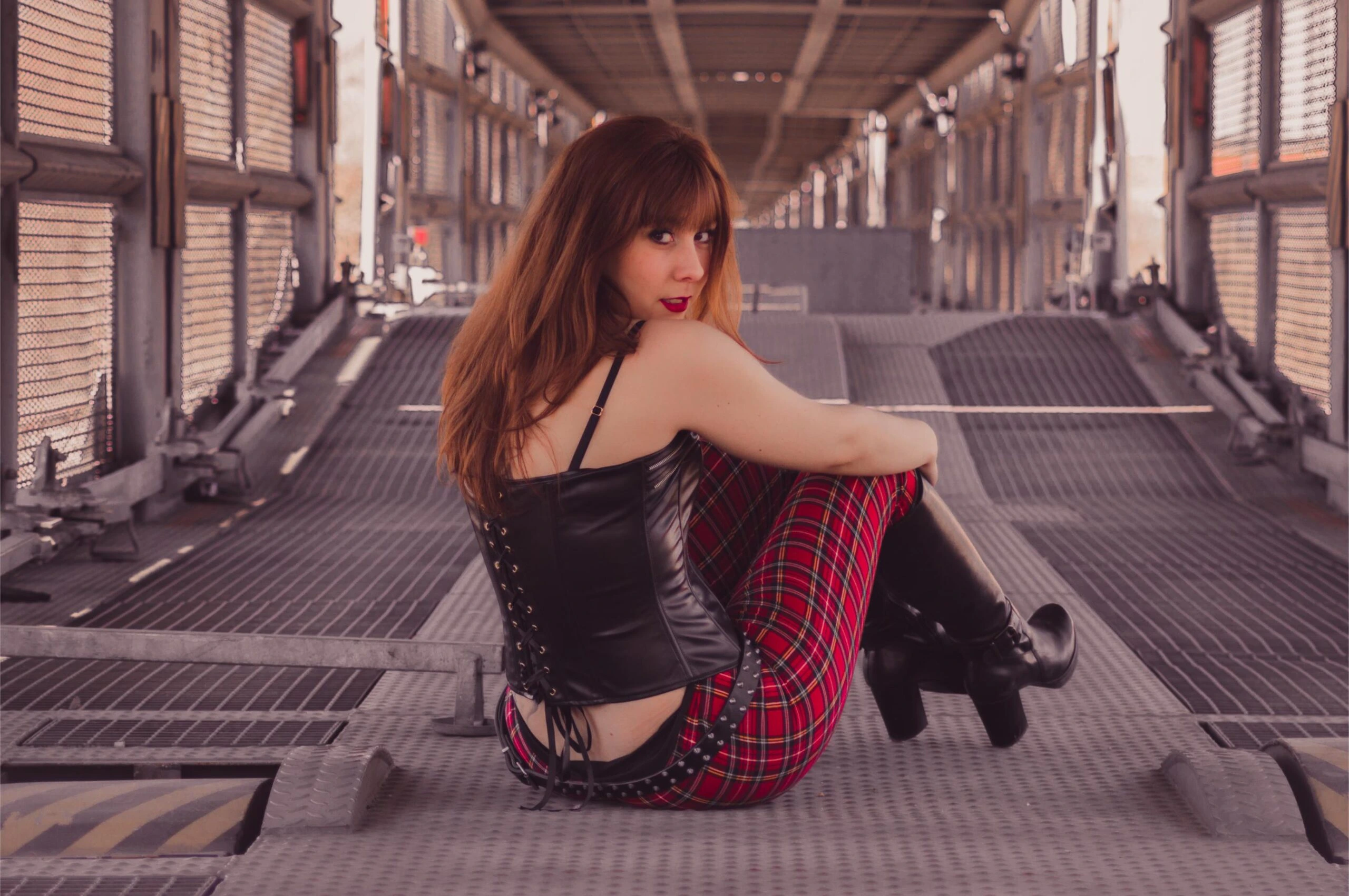 Woman in leather corset and red plaid pants sitting on metal bridge walkway.