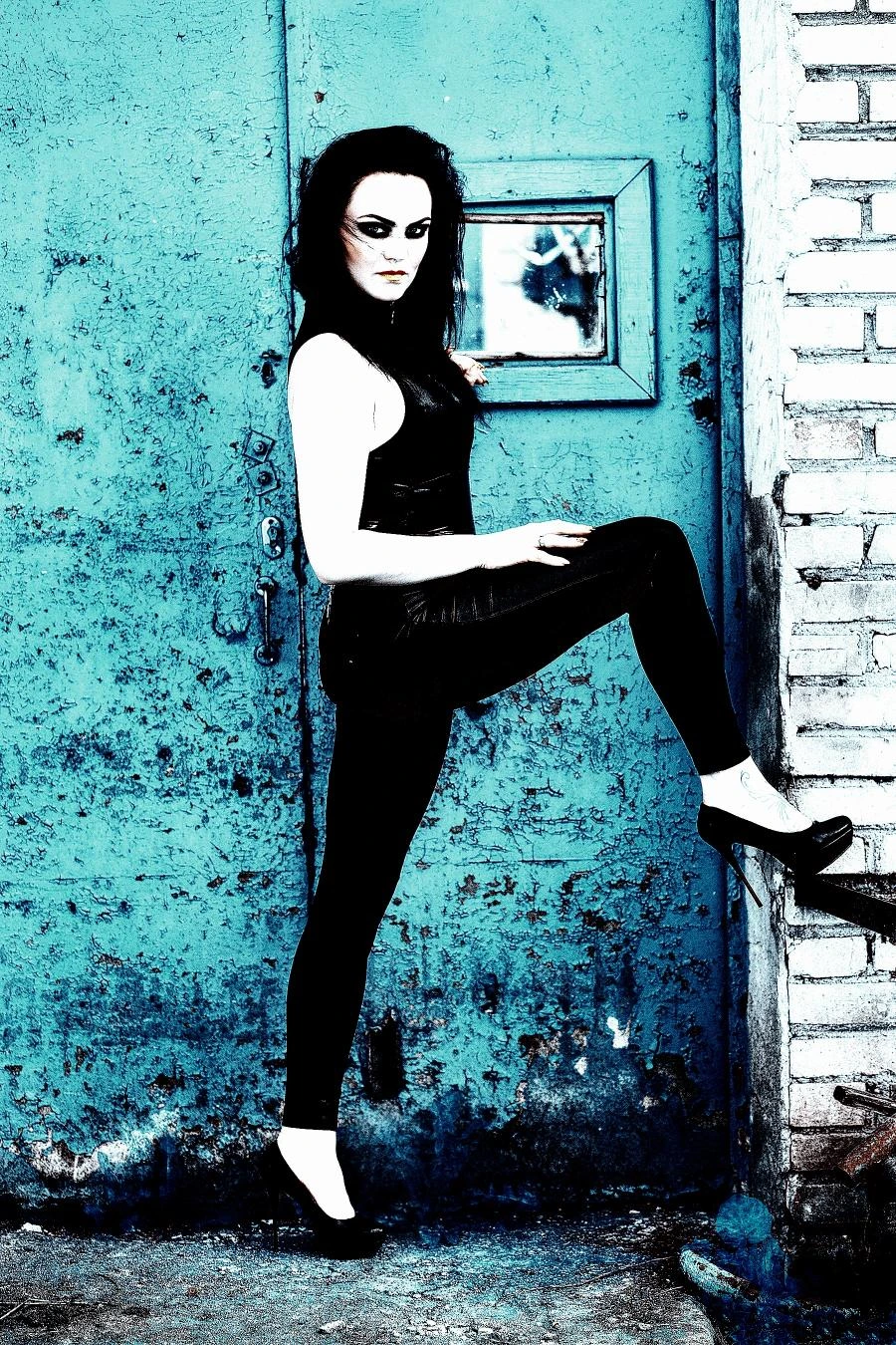Gothic woman in black outfit and heels posing with one foot on step in front of distressed blue metal door. 