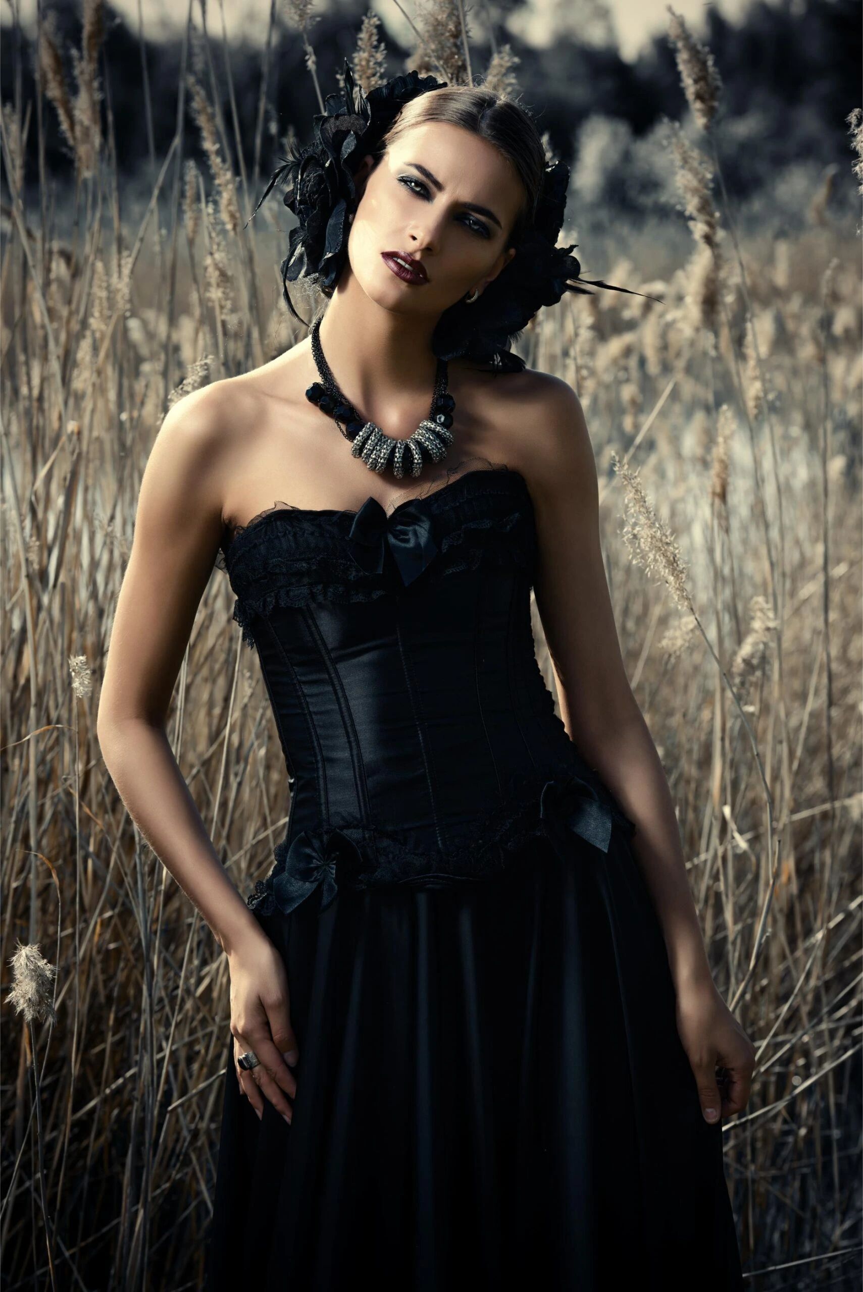 Woman in black corset dress with floral headpiece standing in tall dry grass.